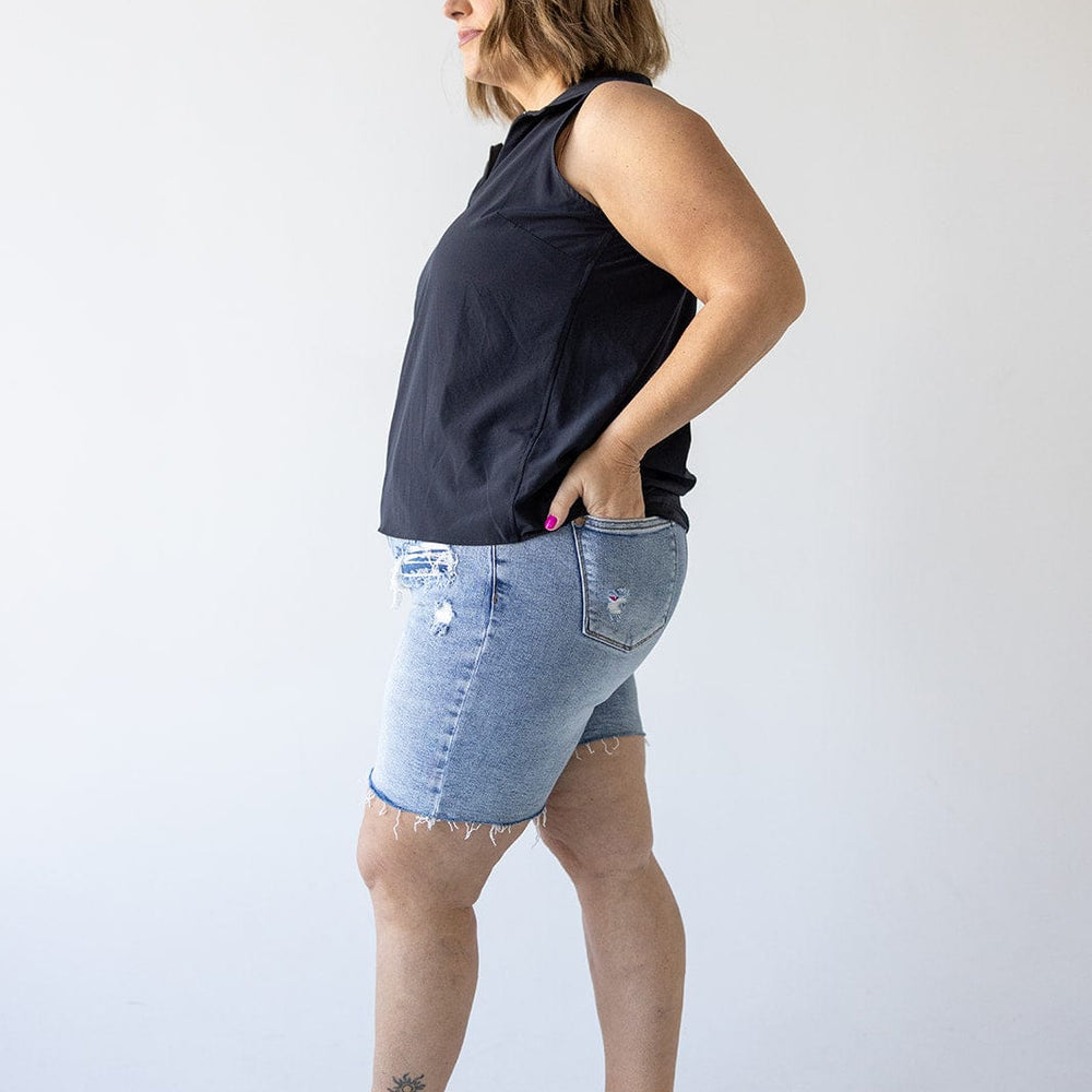 A woman in a sleeveless black top and denim shorts with a high-rise waistline stands side-on against a plain background.