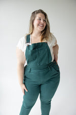 A person with long hair wearing a white shirt and the Judy Blue High-Waisted Double Cuff Overalls in Teal from the brand Judy Blue is standing and facing away from the camera against a plain background.