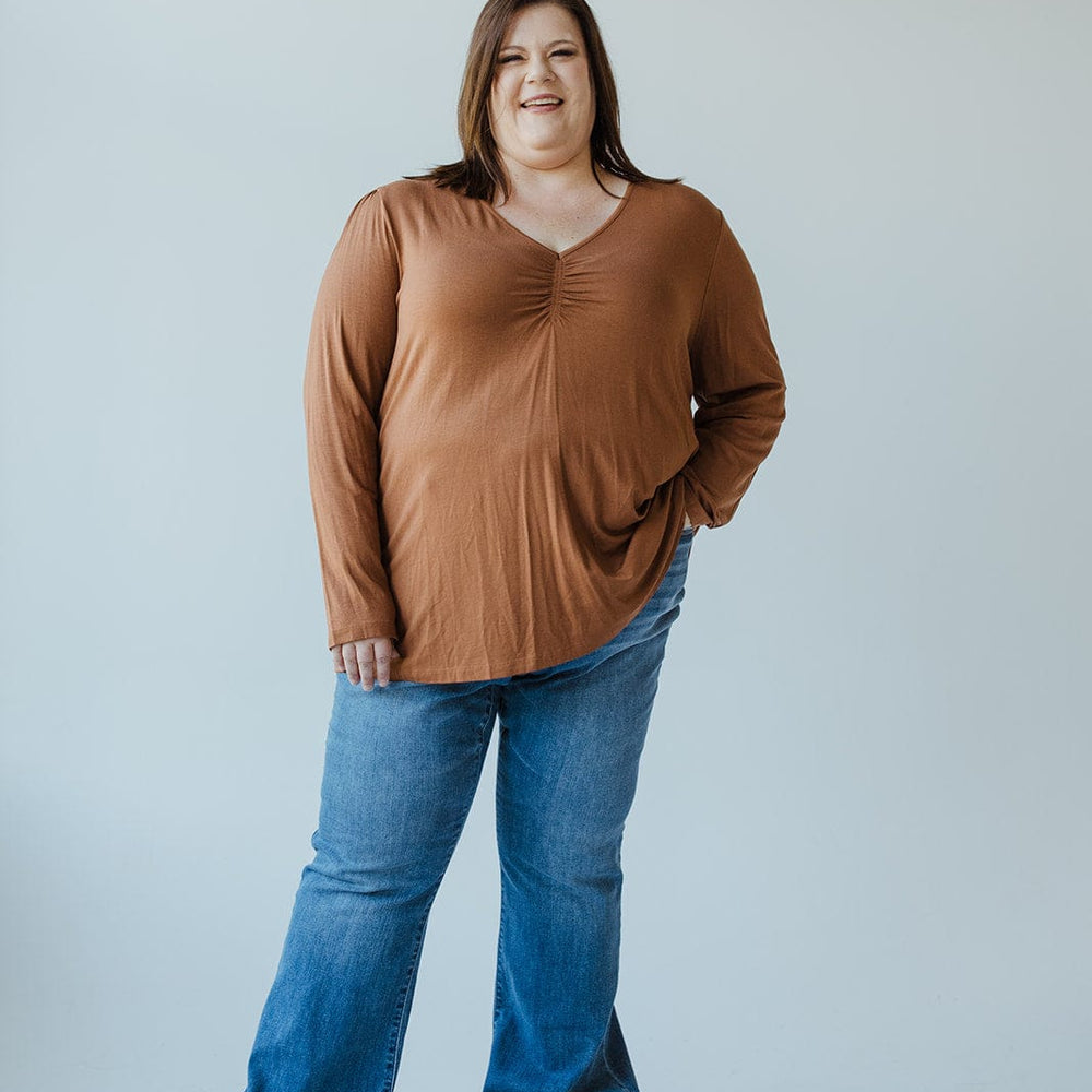 A woman is standing against a plain background, wearing a rust-colored long-sleeve shirt and Judy Blue high-waist non-distressed classic flare jeans in medium blue. She has one hand in her pocket, is smiling, and is also wearing gray shoes.