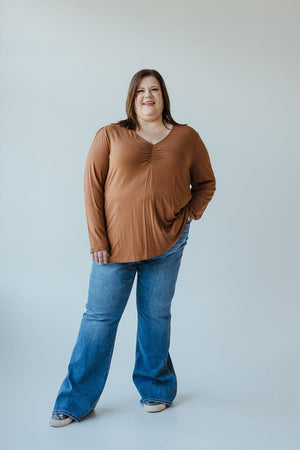 A woman is standing against a plain background, wearing a rust-colored long-sleeve shirt and Judy Blue high-waist non-distressed classic flare jeans in medium blue. She has one hand in her pocket, is smiling, and is also wearing gray shoes.