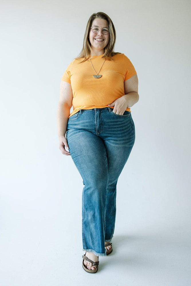 A person in an orange shirt and Judy Blue high-waist tummy control flare jeans with a frayed hem stands against a plain background, smiling and holding a necklace.