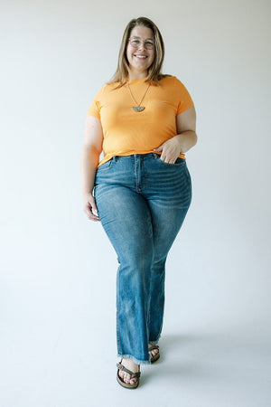 A person in an orange shirt and Judy Blue high-waist tummy control flare jeans with a frayed hem stands against a plain background, smiling and holding a necklace.