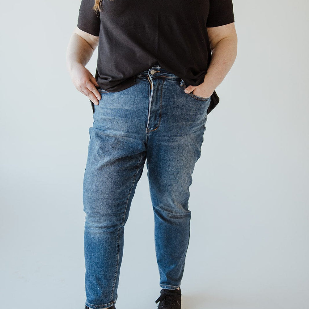 A person stands against a plain background, wearing a black V-neck shirt, Judy Blue High Rise Tummy Control Slim Straight Jeans by Judy Blue, and black sneakers with white soles. Their hands are in their pockets.