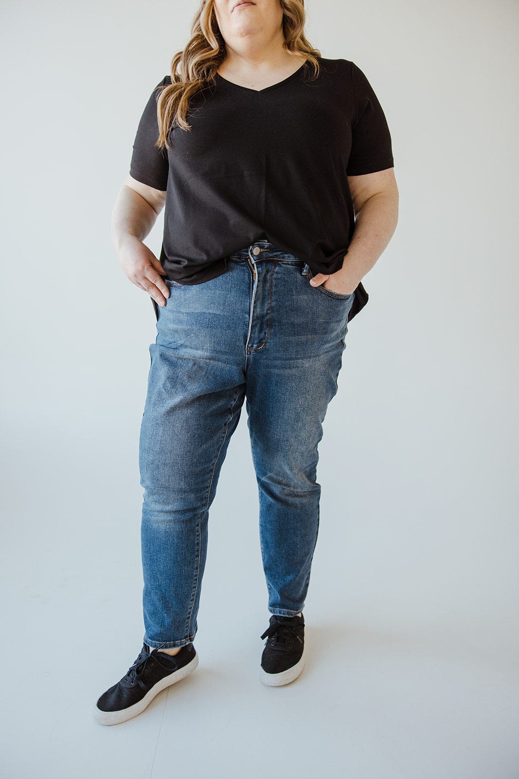 A person stands against a plain background, wearing a black V-neck shirt, Judy Blue High Rise Tummy Control Slim Straight Jeans by Judy Blue, and black sneakers with white soles. Their hands are in their pockets.