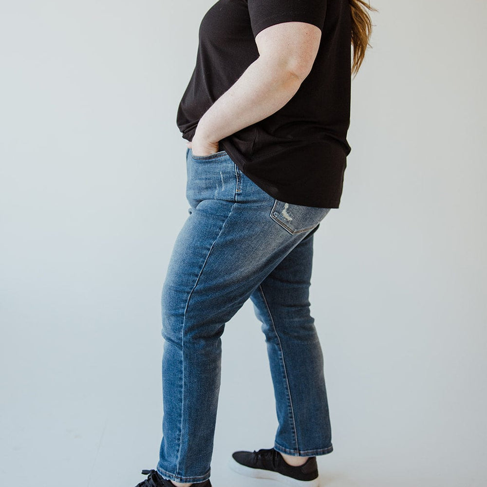 A person wearing a black t-shirt, black sneakers, and Judy Blue High Rise Tummy Control Slim Straight Jeans with light distressing on the pockets stands with hands in their pockets and looks to the side.