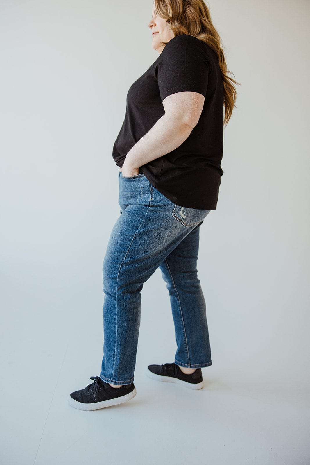 A person wearing a black t-shirt, black sneakers, and Judy Blue High Rise Tummy Control Slim Straight Jeans with light distressing on the pockets stands with hands in their pockets and looks to the side.