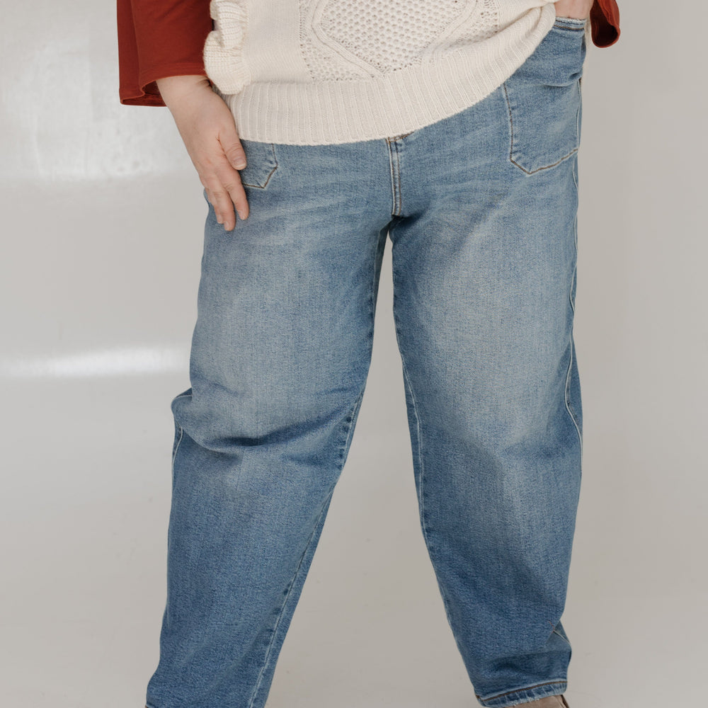 Person wearing a beige sweater, red shirt, blue jeans, and brown shoes on a white background
