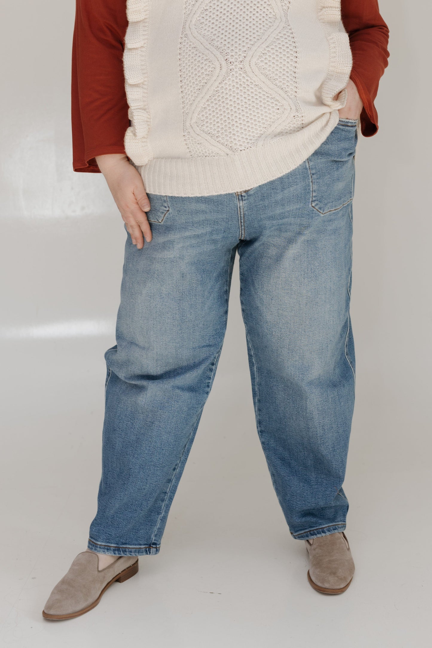 Person wearing a beige sweater, red shirt, blue jeans, and brown shoes on a white background