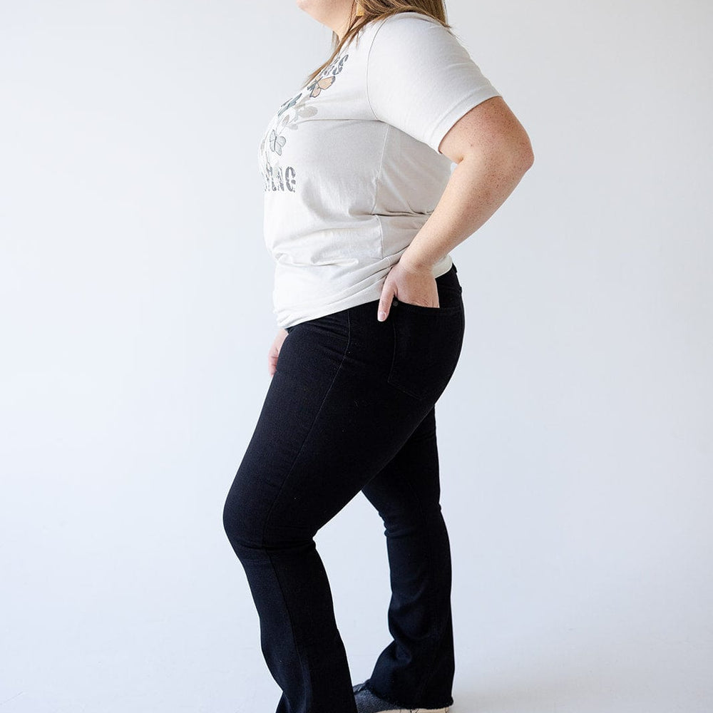 A person stands in profile view, wearing a light-colored T-shirt and black button fly jeans with one hand in a pocket, against a plain white background.