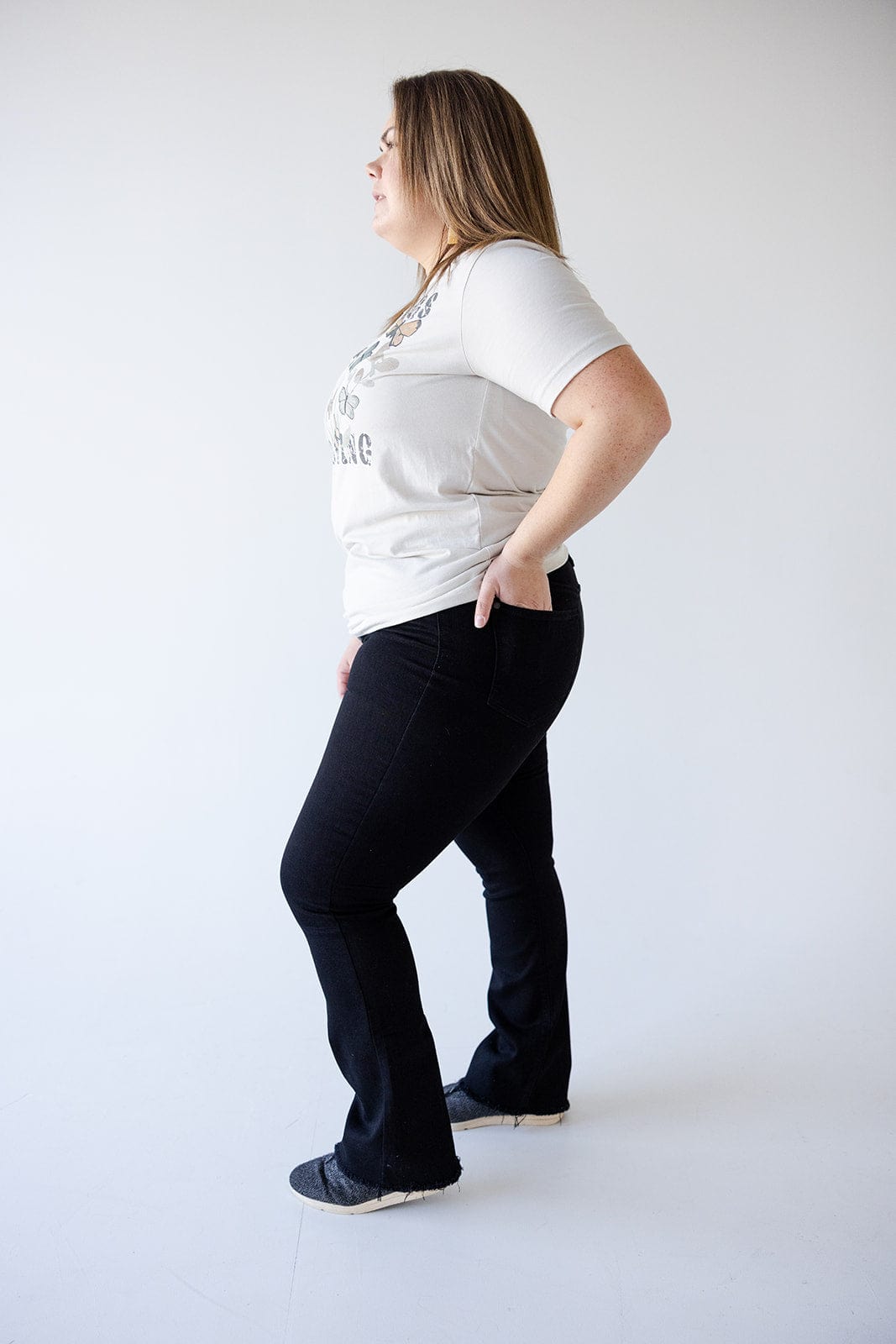 A person stands in profile view, wearing a light-colored T-shirt and black button fly jeans with one hand in a pocket, against a plain white background.