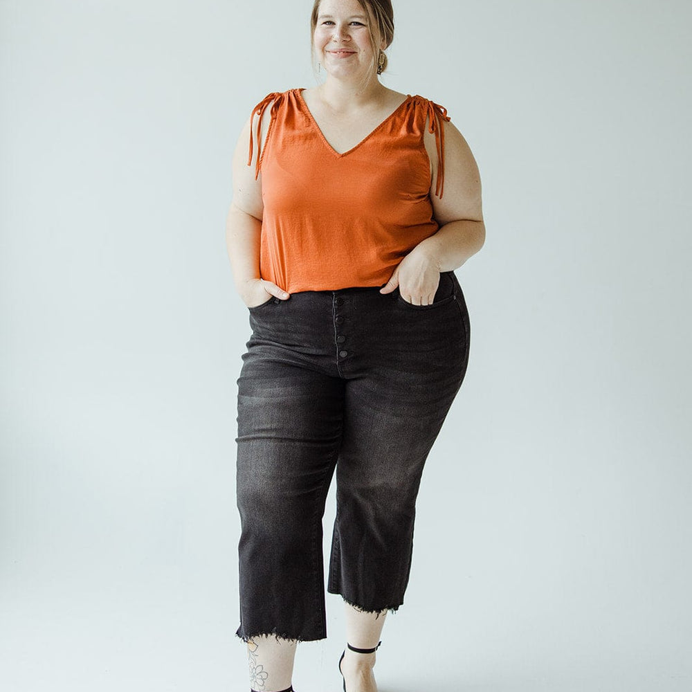 A woman stands against a plain background, wearing an orange sleeveless top and Judy Blue JUDY BLUE HIGH WAIST BUTTON FLY RAW HEM CROPPED WIDE LEG jeans. She completes the look with black high-heeled sandals, has one hand in her pocket, and is smiling.