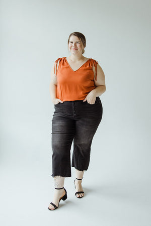 A woman stands against a plain background, wearing an orange sleeveless top and Judy Blue JUDY BLUE HIGH WAIST BUTTON FLY RAW HEM CROPPED WIDE LEG jeans. She completes the look with black high-heeled sandals, has one hand in her pocket, and is smiling.