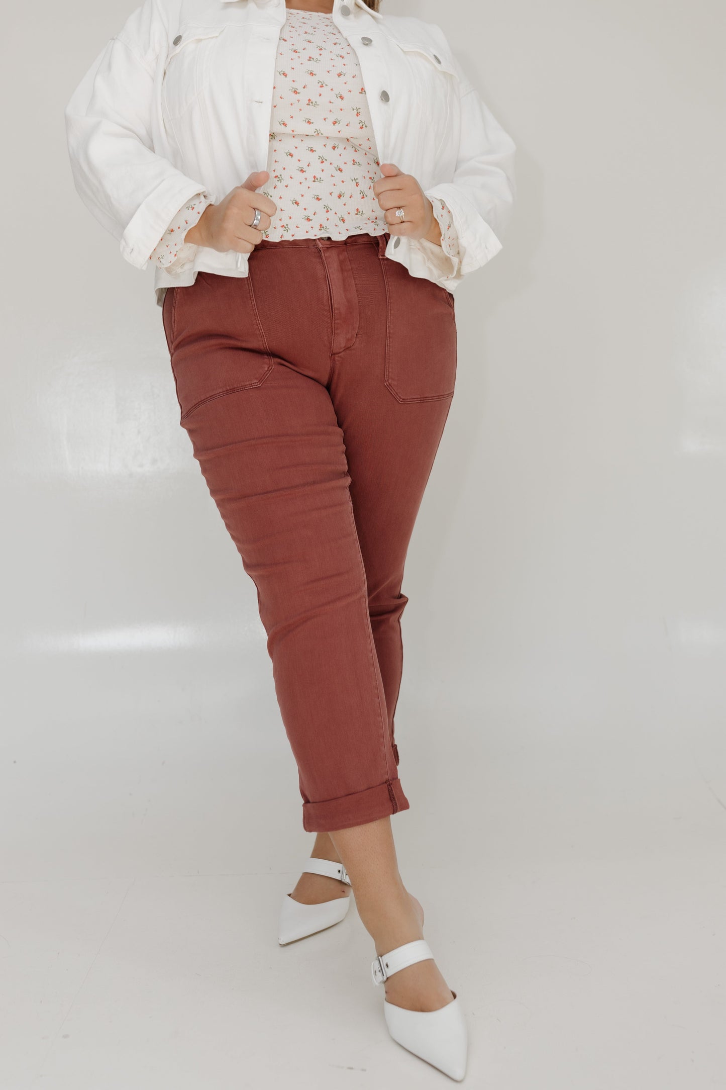 Person wearing a white jacket, patterned top, rust-colored pants, and white shoes on a light gray background