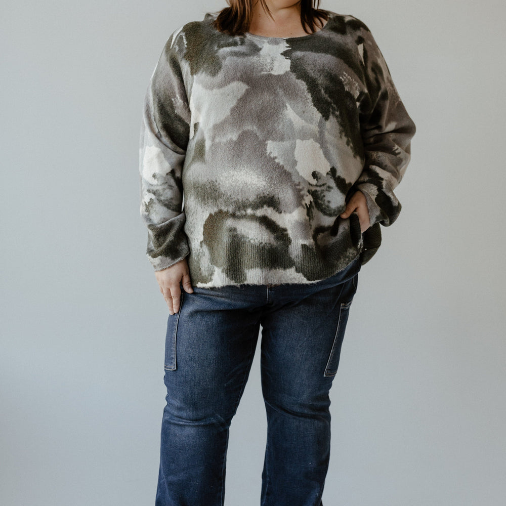 A person stands against a neutral background, wearing a camouflage-patterned sweater and Judy Blue High Waist Dark Wash Bootcut jeans with cargo pockets, with one hand in their pocket.