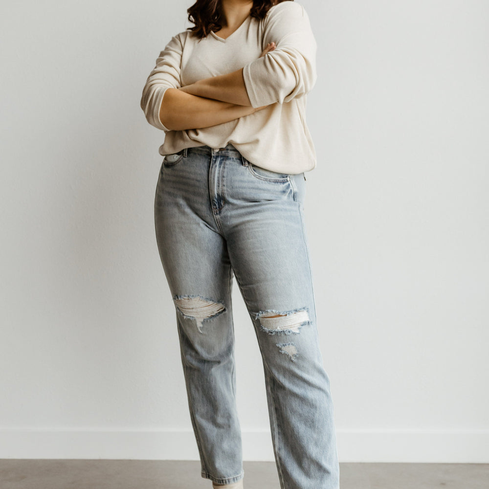 A person stands with arms crossed, wearing a beige sweater, Judy Blue high waist jeans with a destroyed 90's style, and white boots, set against a plain white background.