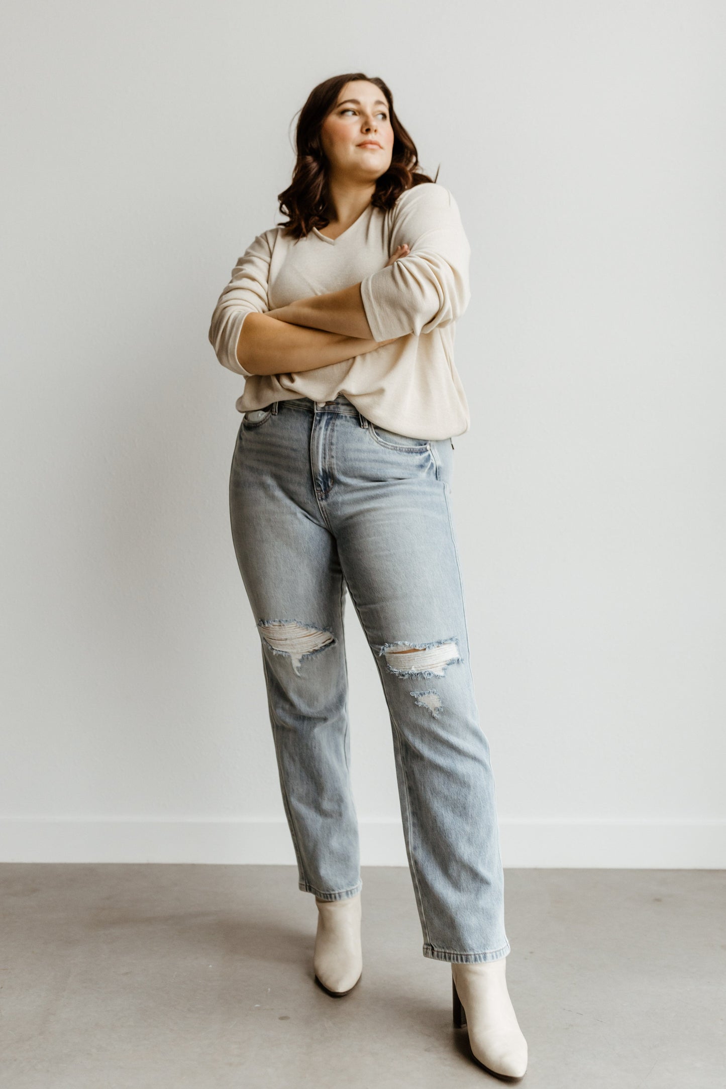 A person stands with arms crossed, wearing a beige sweater, Judy Blue high waist jeans with a destroyed 90's style, and white boots, set against a plain white background.