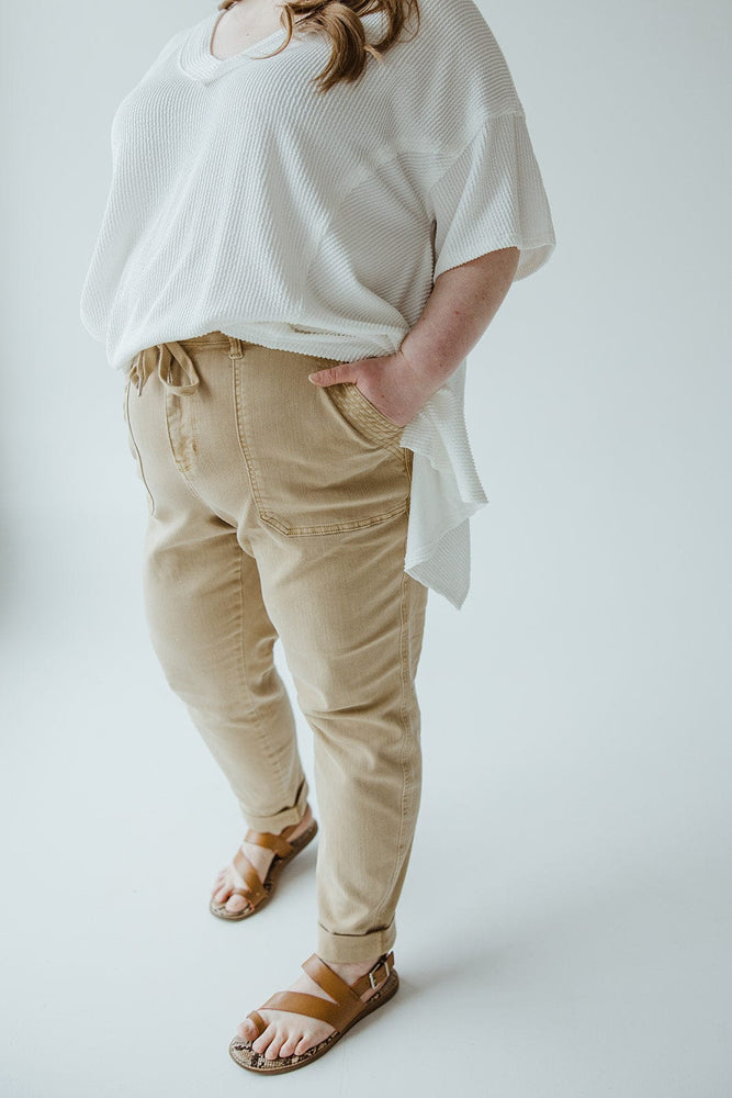 Person stands with one hand in pocket, wearing a loose white top, Judy Blue JUDY BLUE HIGH WAIST DOUBLE ROLLED CUFFED JOGGER JEANS IN KHAKI, and brown sandals, against a plain white background.