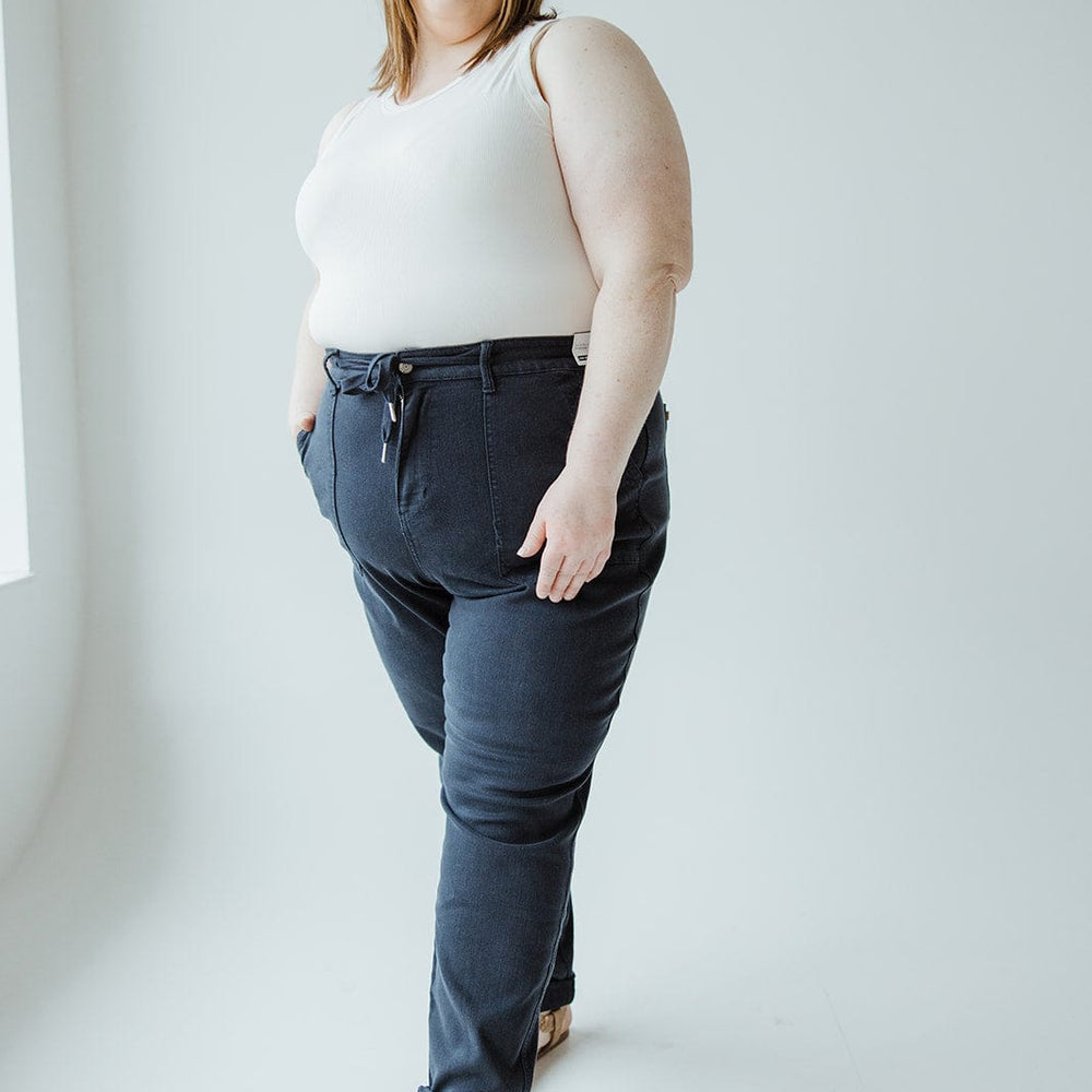 A woman stands against a light background, wearing a white tank top, Judy Blue High Waist Double Rolled Cuffed Jogger Jeans in Navy by Judy Blue, and sandals, with her hands in her pockets, looking at the camera.