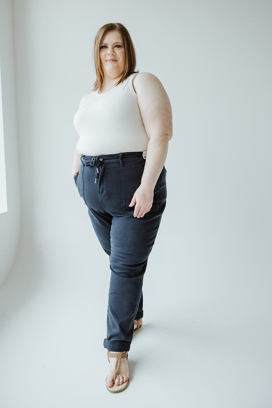 A woman stands against a light background, wearing a white tank top, Judy Blue High Waist Double Rolled Cuffed Jogger Jeans in Navy by Judy Blue, and sandals, with her hands in her pockets, looking at the camera.