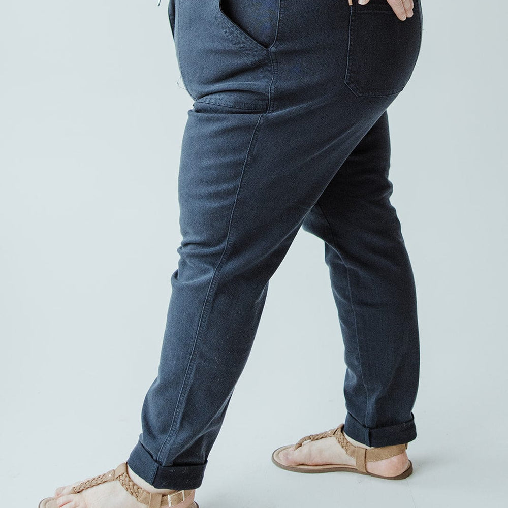 A person wearing a white top, Judy Blue JUDY BLUE HIGH WAIST DOUBLE ROLLED CUFFED JOGGER JEANS IN NAVY, and tan sandals stands with one hand in their pocket against a light background.