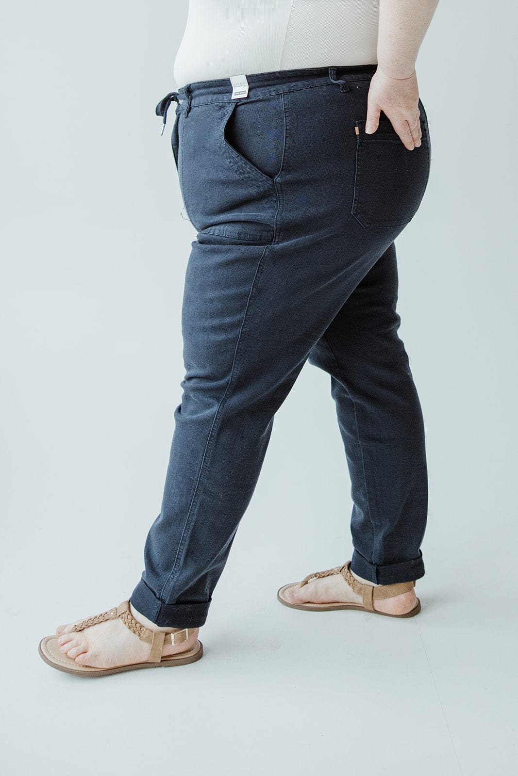 A person wearing a white top, Judy Blue JUDY BLUE HIGH WAIST DOUBLE ROLLED CUFFED JOGGER JEANS IN NAVY, and tan sandals stands with one hand in their pocket against a light background.