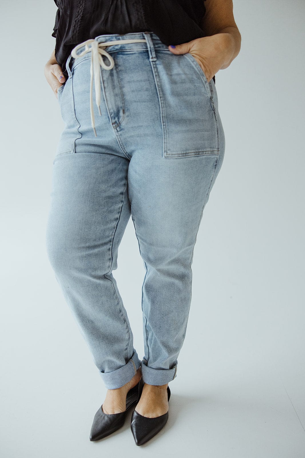 A person wearing light blue Judy Blue high-waisted jeans, with hands in pockets, and a black top. The ultra-stretchy denim adds comfort to their look, complemented by black pointed shoes.