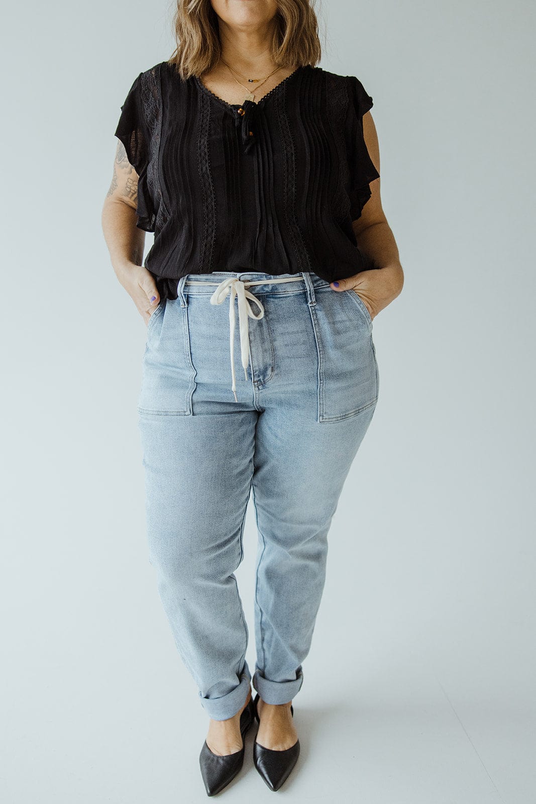 A person wearing a black blouse, light blue comfort jeans with a drawstring, and black shoes stands against a plain background.