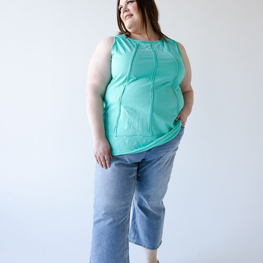 A person in a turquoise sleeveless top and wide leg crop jeans stands against a plain background, looking to their left with a hand in one pocket.