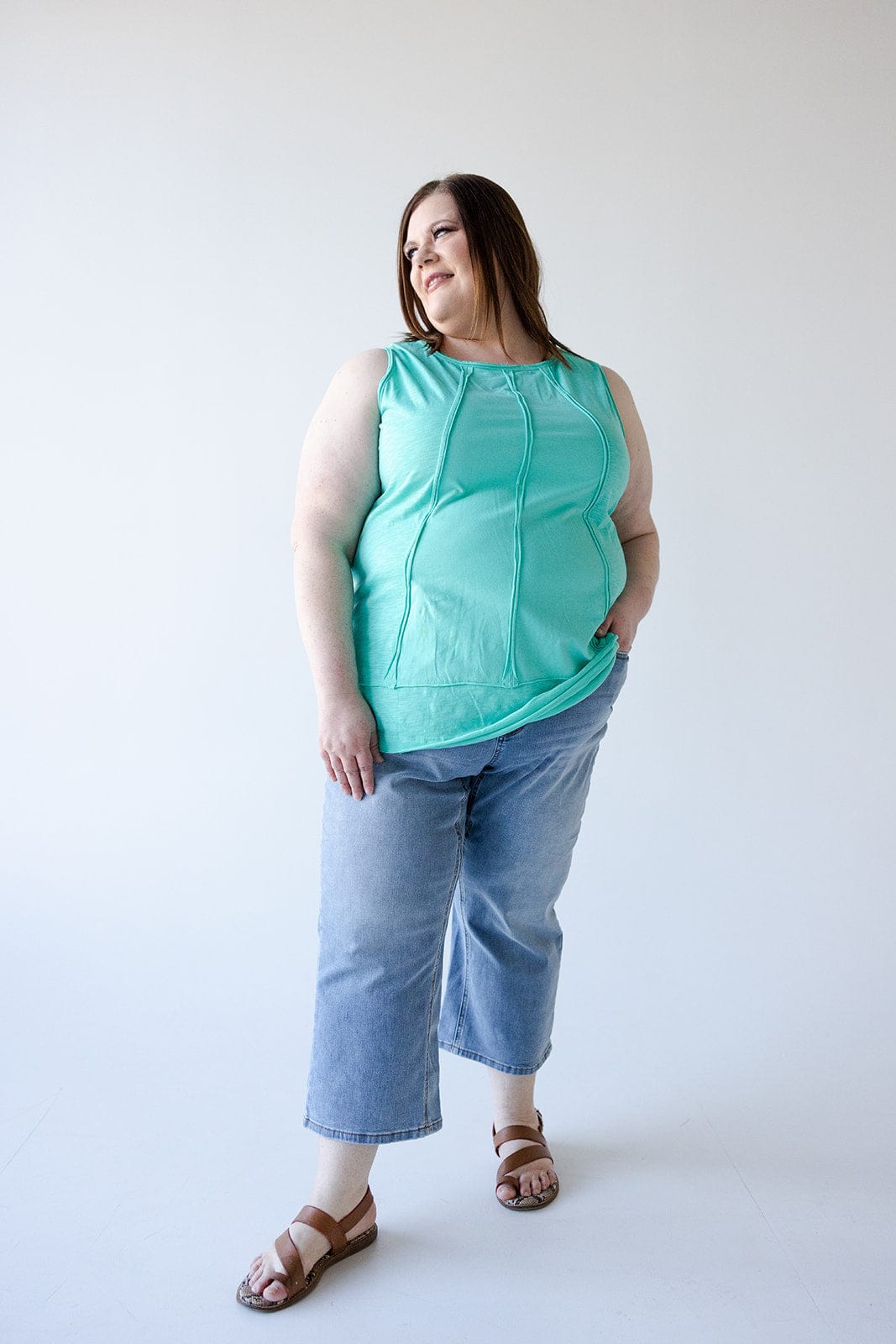 A person in a turquoise sleeveless top and wide leg crop jeans stands against a plain background, looking to their left with a hand in one pocket.