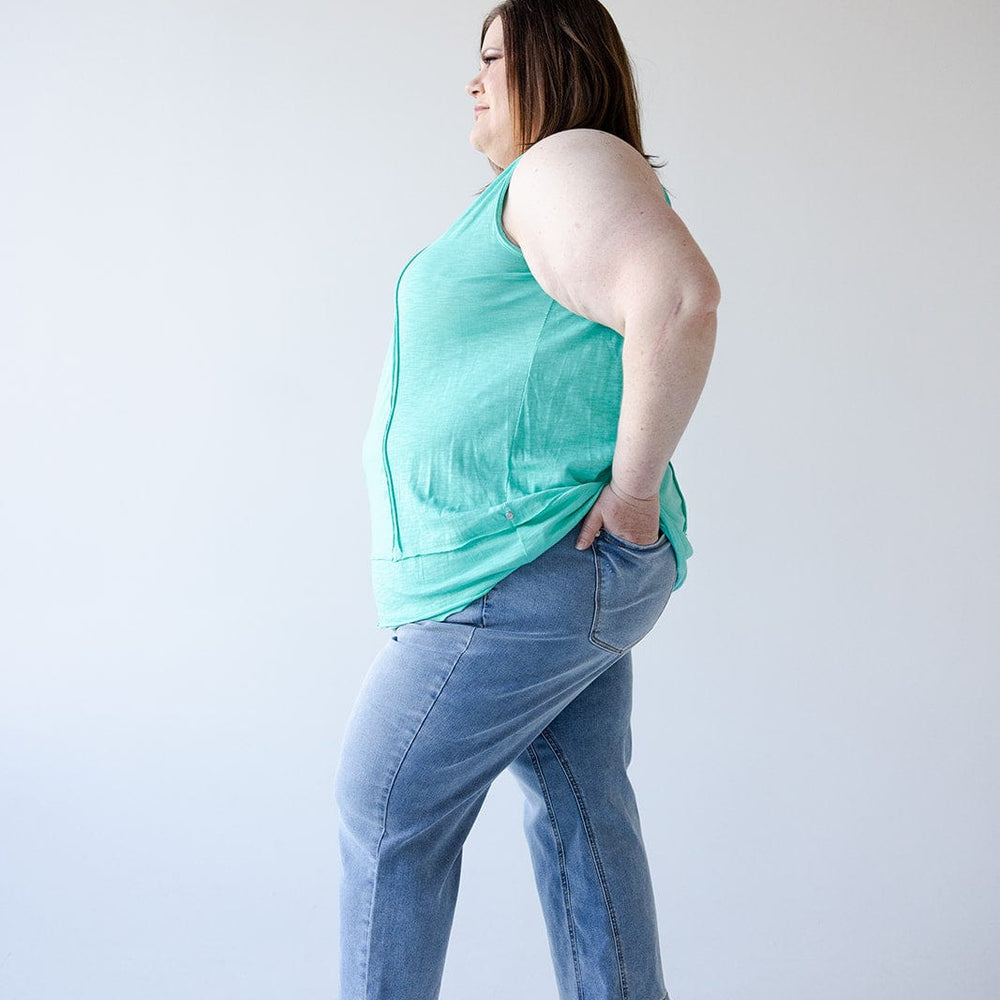 A person in a turquoise tank top and jeans featuring a high waist double waistband stands sideways with hands in pockets against a plain background.