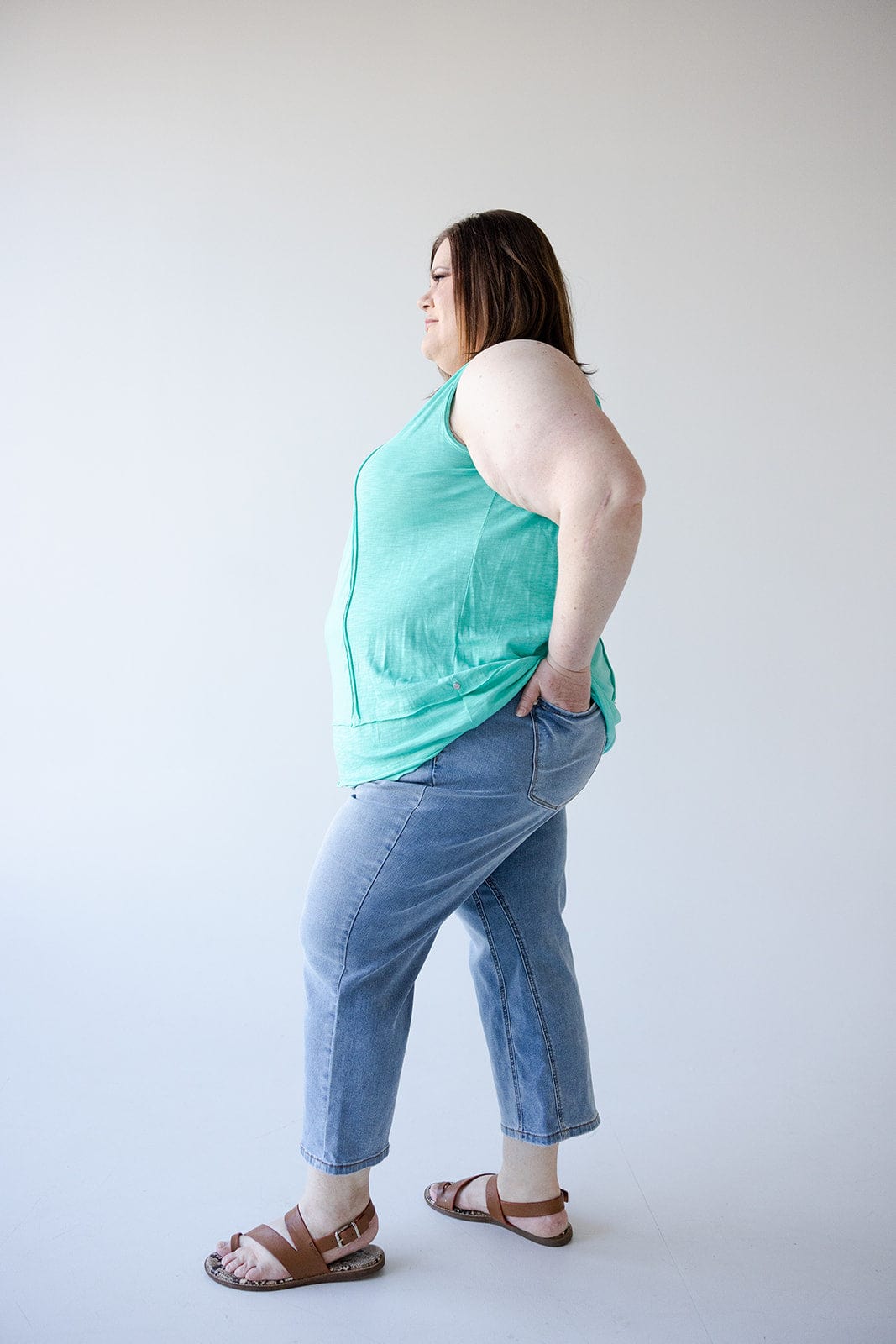 A person in a turquoise tank top and jeans featuring a high waist double waistband stands sideways with hands in pockets against a plain background.