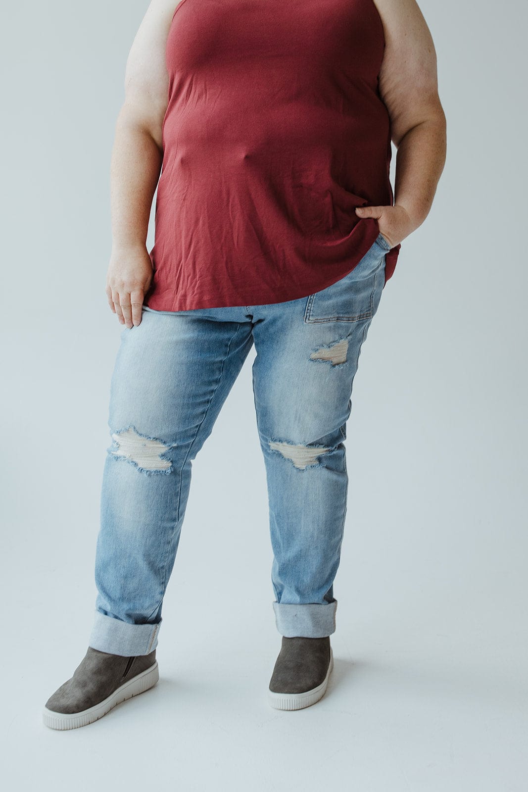 A person rocking the plus-size cool girl look, wearing a red sleeveless top, Judy Blue High Waisted Destroyed Boyfriend Jeans in Medium Wash made from Judy Blue’s stretchy denim, and gray high-top sneakers with one hand in their pocket.