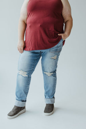 A person rocking the plus-size cool girl look, wearing a red sleeveless top, Judy Blue High Waisted Destroyed Boyfriend Jeans in Medium Wash made from Judy Blue’s stretchy denim, and gray high-top sneakers with one hand in their pocket.