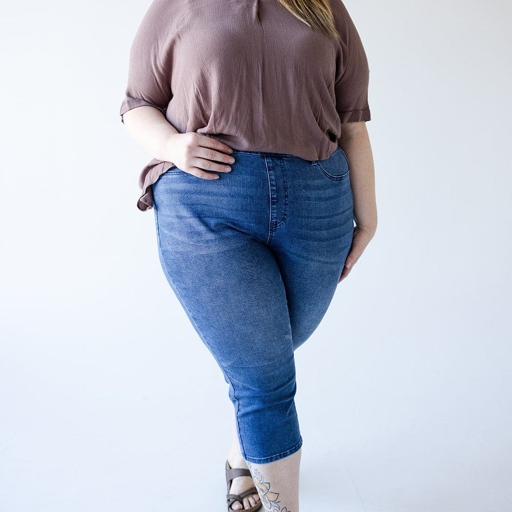 A person stands smiling, wearing a brown top, Judy Blue High-Waisted Pull-On Capri with Cool Denim Technology in Medium Wash, and sandals. They have glasses and a tattoo on their lower leg.