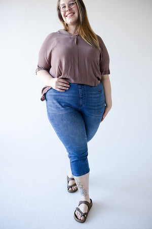 A person stands smiling, wearing a brown top, Judy Blue High-Waisted Pull-On Capri with Cool Denim Technology in Medium Wash, and sandals. They have glasses and a tattoo on their lower leg.