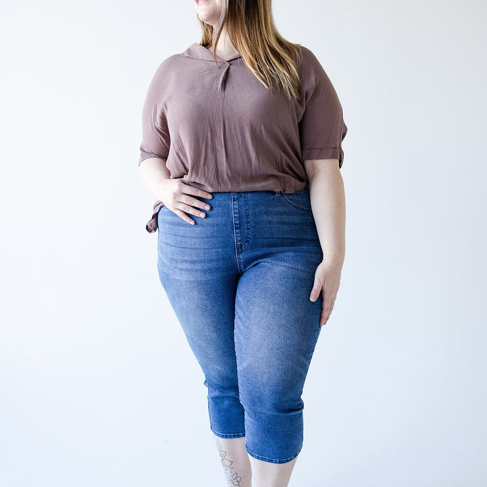 A person is standing, wearing a brown top, Judy Blue High Waisted Pull-On Capri with Cool Denim Technology in Medium Wash, and sandals with glasses. They have long hair with partial highlights and are smiling with one hand in their pocket.