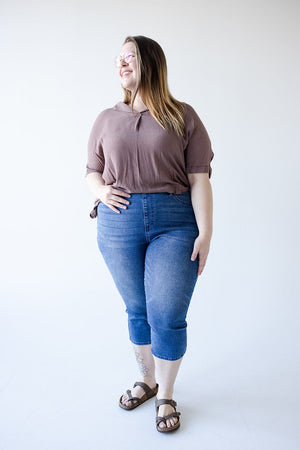 A person is standing, wearing a brown top, Judy Blue High Waisted Pull-On Capri with Cool Denim Technology in Medium Wash, and sandals with glasses. They have long hair with partial highlights and are smiling with one hand in their pocket.