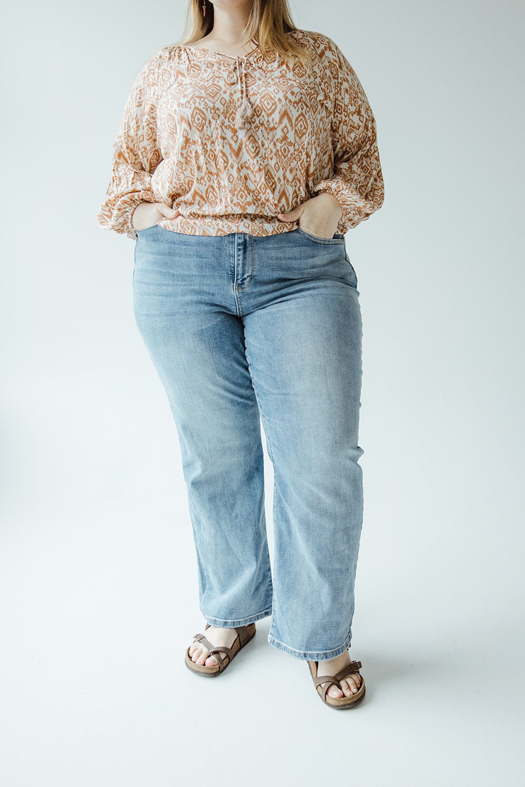 Person wearing a patterned top, JUDY BLUE HIGH WAIST NON-DISTRESSED STRAIGHT LEG JEAN by Judy Blue, and sandals stands with hands in pockets against a plain background.