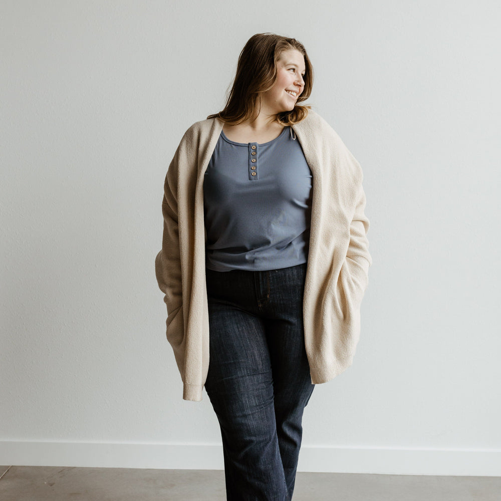 A person stands against a plain wall, wearing a light cardigan, blue top, and Judy Blue Jeans, looking to the side with a relaxed expression.