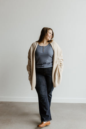 A person stands against a plain wall, wearing a light cardigan, blue top, and Judy Blue Jeans, looking to the side with a relaxed expression.