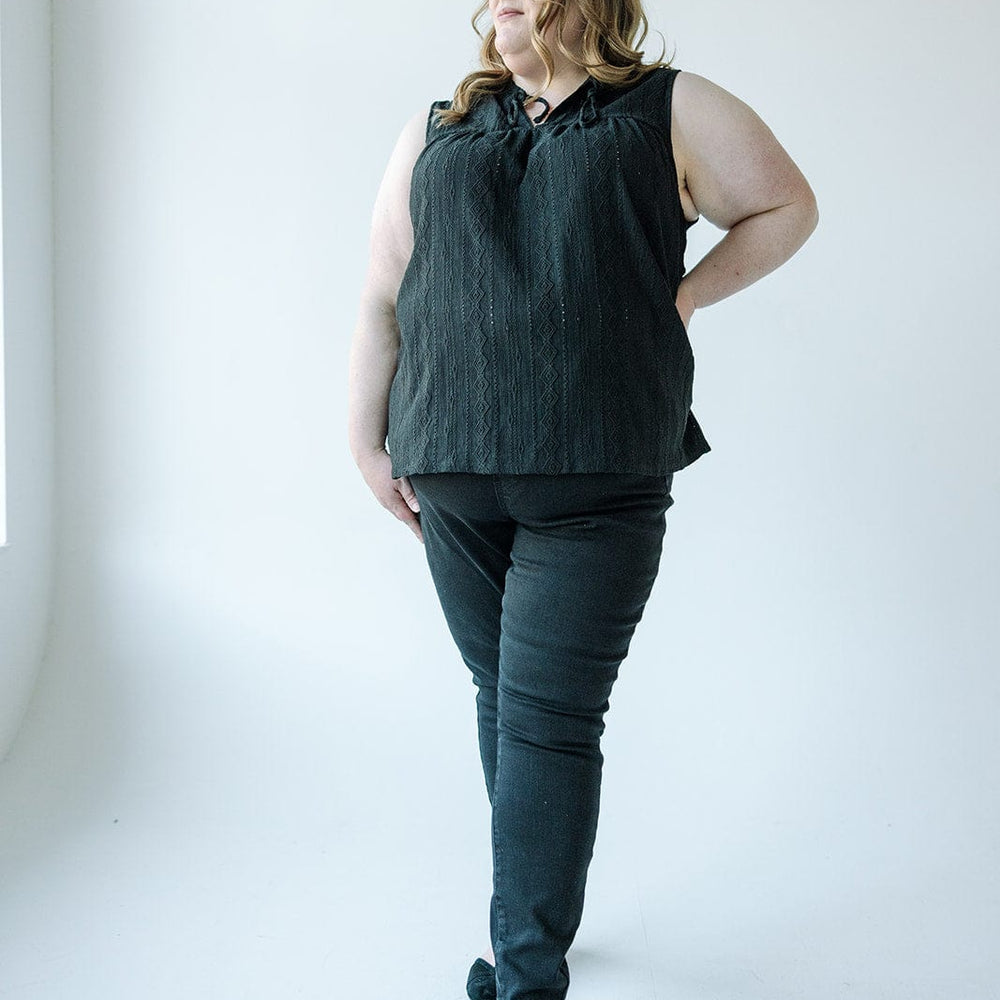 A person with long hair stands confidently in front of a light-colored wall, wearing a sleeveless black top, Judy Blue High Waist Pull-On Skinny Jeans in Black, and black shoes.