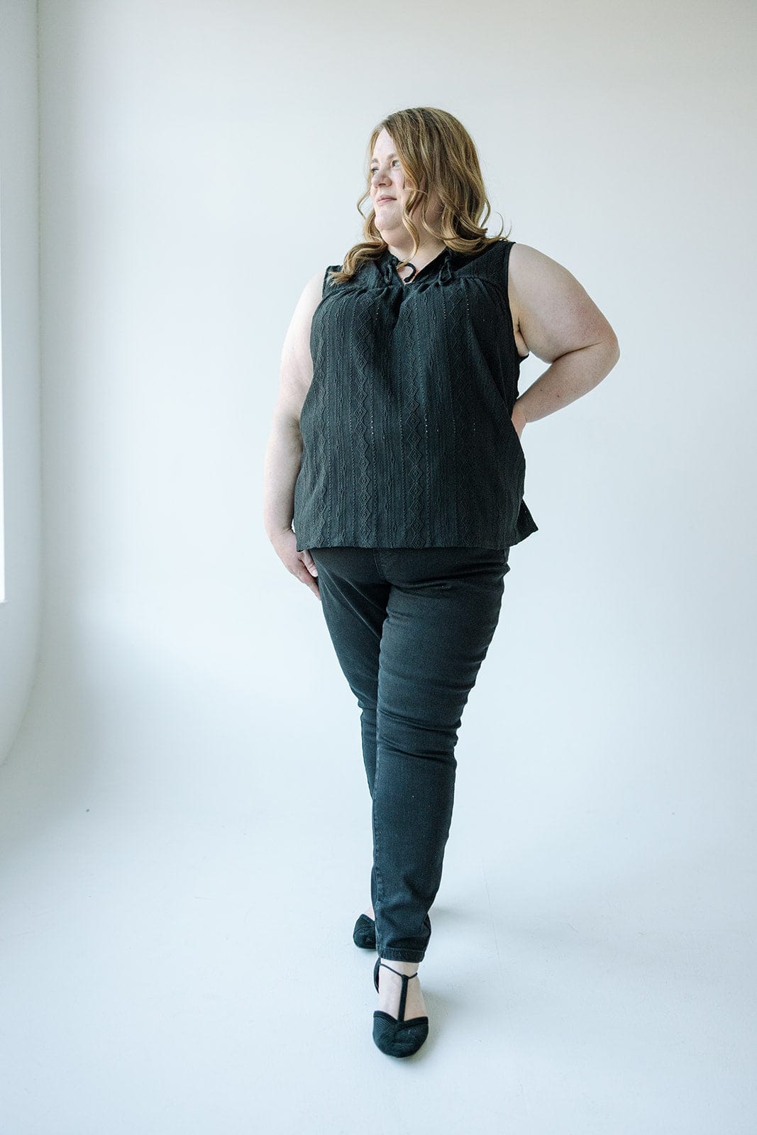 A person with long hair stands confidently in front of a light-colored wall, wearing a sleeveless black top, Judy Blue High Waist Pull-On Skinny Jeans in Black, and black shoes.