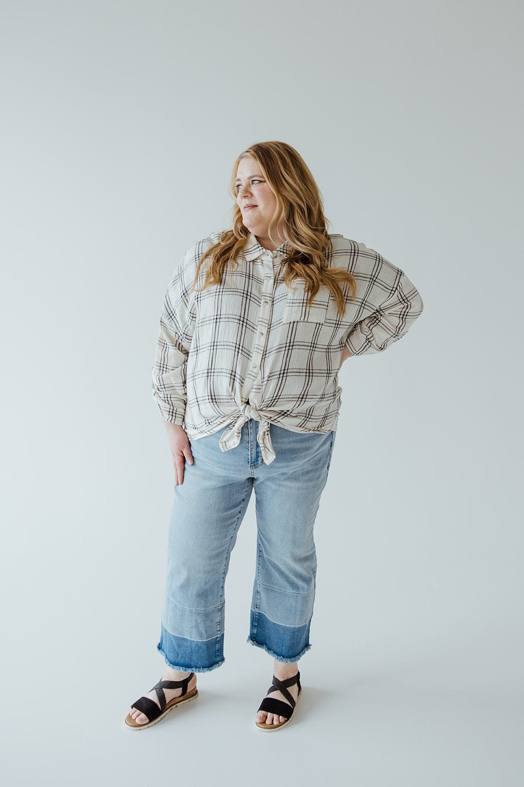 A woman stands with one hand on her hip, wearing a plaid shirt, Judy Blue JUDY BLUE HIGH WAIST RELEASED HEM CROPPED WIDE LEG jeans with rolled cuffs, and black sandals, against a plain background.