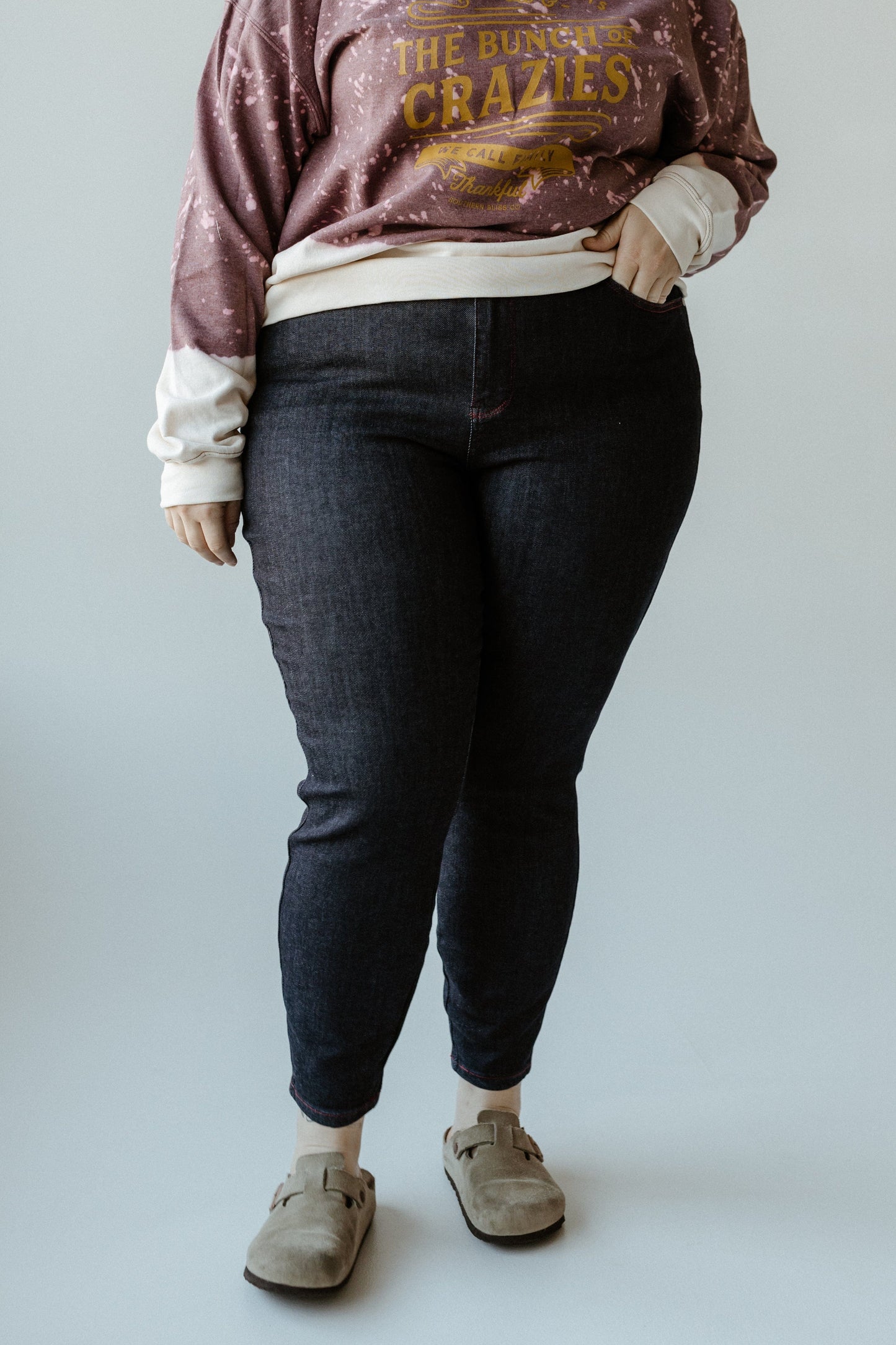 A person wearing a purple sweatshirt that reads "The Bunch of Crazies" pairs it with dark denim high-waist skinny jeans, featuring heart-shaped back pockets, and beige shoes. They stand confidently against a plain background, showcasing their unique style.