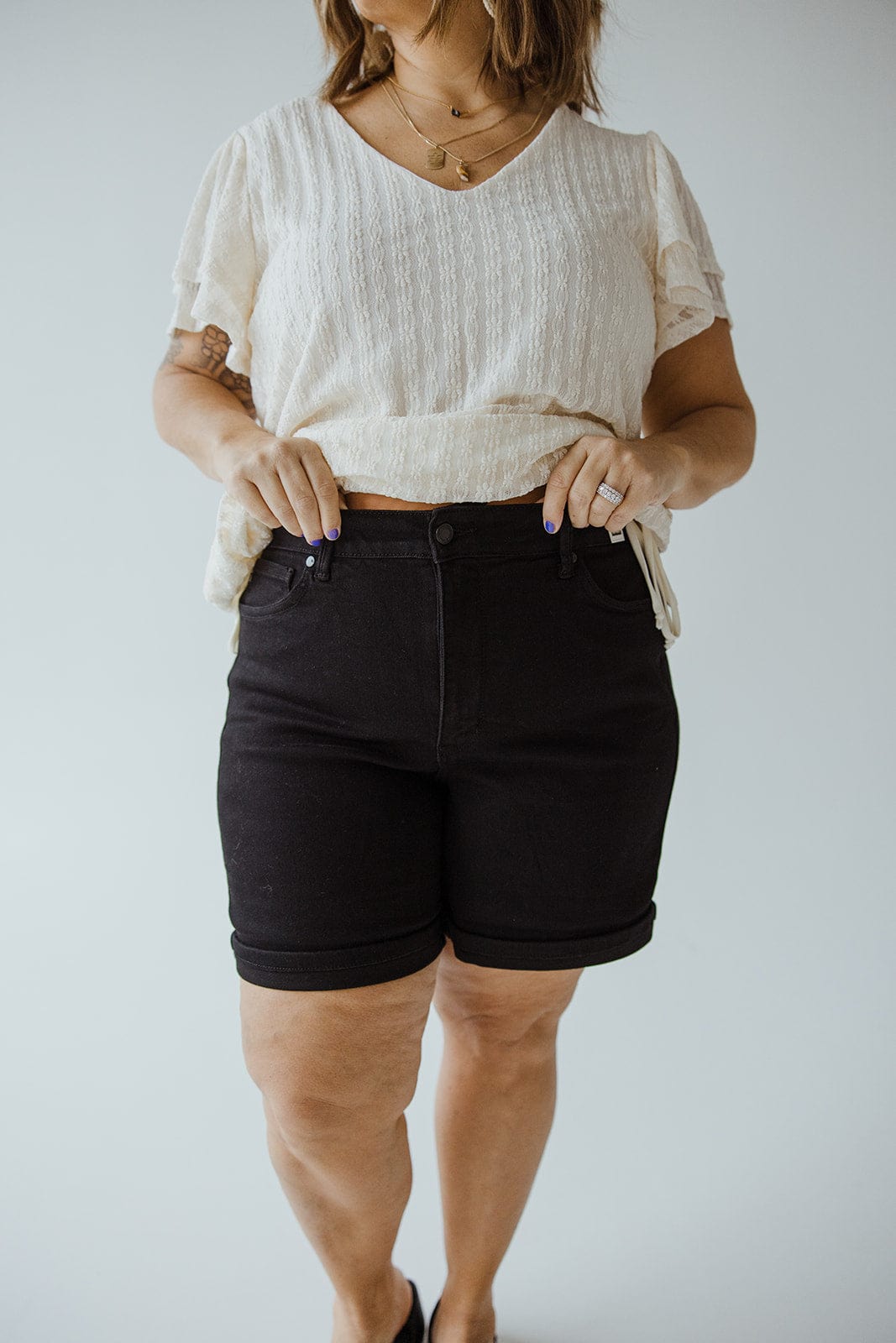 A person wearing a white blouse and Judy Blue JUDY BLUE HIGH WAIST TUMMY CONTROL CUFFED SHORTS IN BLACK poses against a plain background.