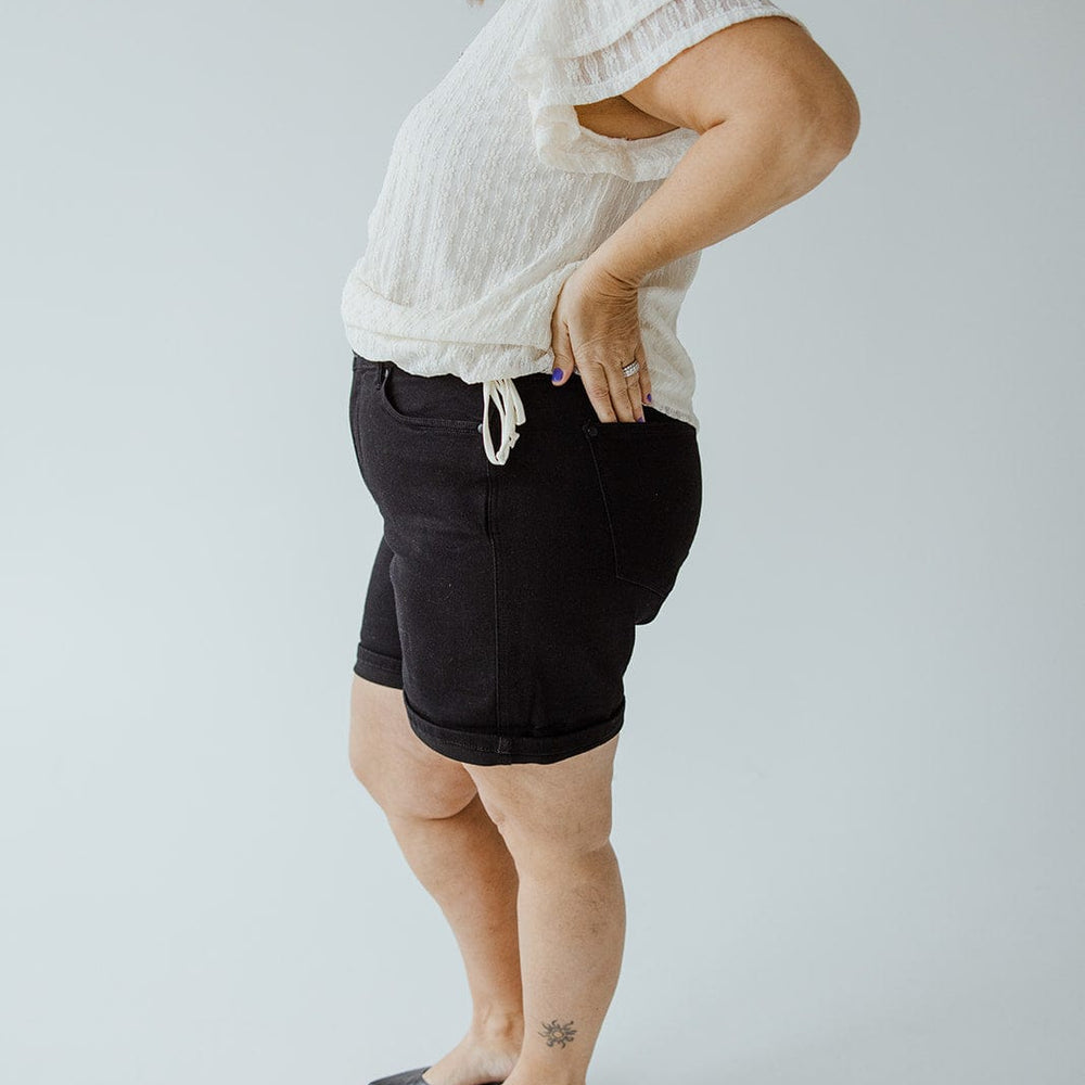 A person standing sideways against a plain background, wearing a white blouse and JUDY BLUE HIGH WAIST TUMMY CONTROL CUFFED SHORTS IN BLACK from Judy Blue, with their hands on their hips.