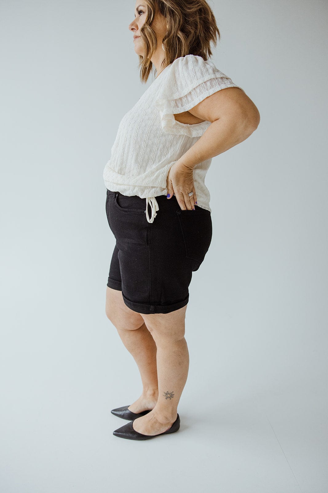 A person standing sideways against a plain background, wearing a white blouse and JUDY BLUE HIGH WAIST TUMMY CONTROL CUFFED SHORTS IN BLACK from Judy Blue, with their hands on their hips.