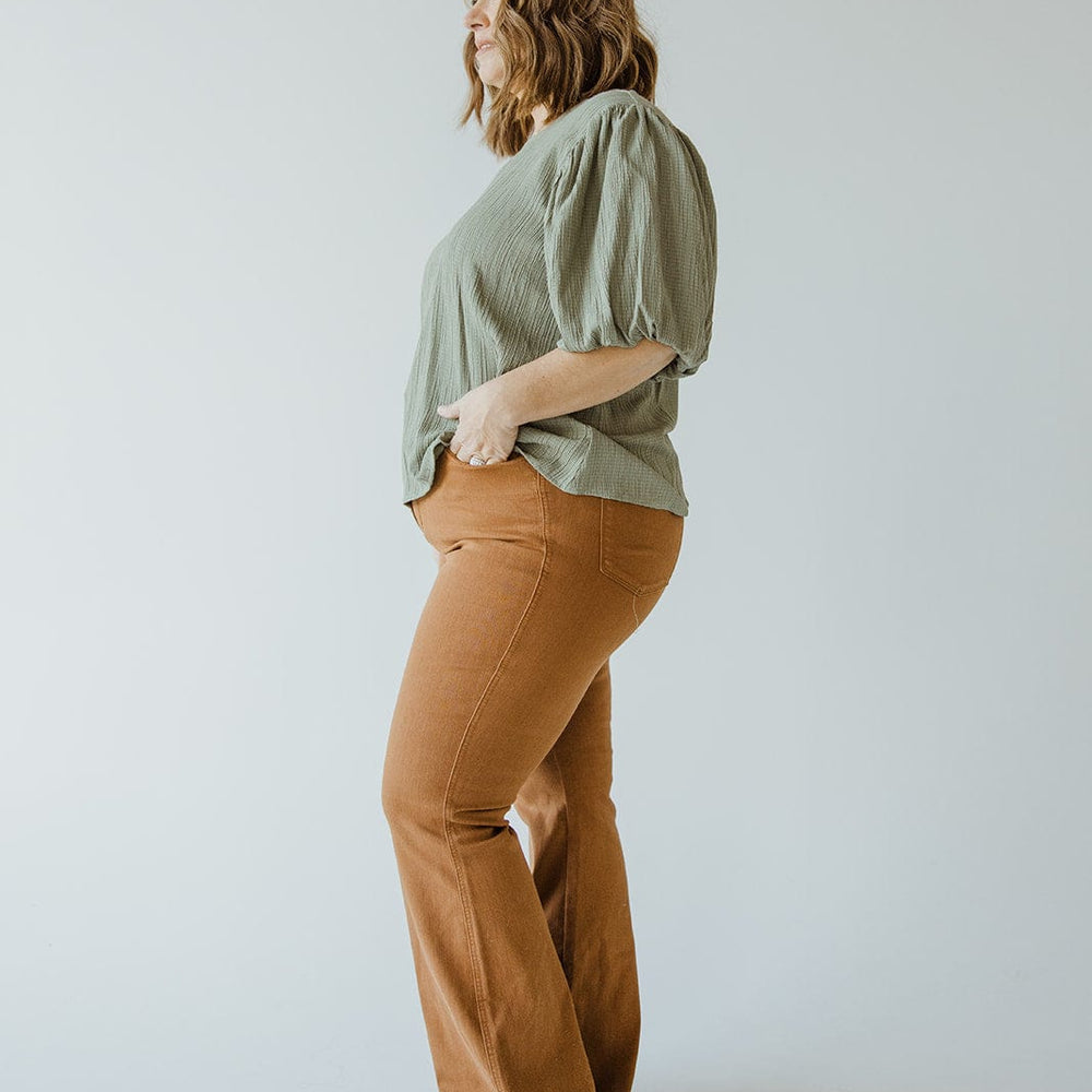 A woman stands sideways, wearing a green blouse with puffed sleeves and JUDY BLUE HIGH WAIST TUMMY CONTROL FLARE IN URBAN BRONZE pants by Judy Blue. She looks away with her hands in her pockets against a plain, light background.