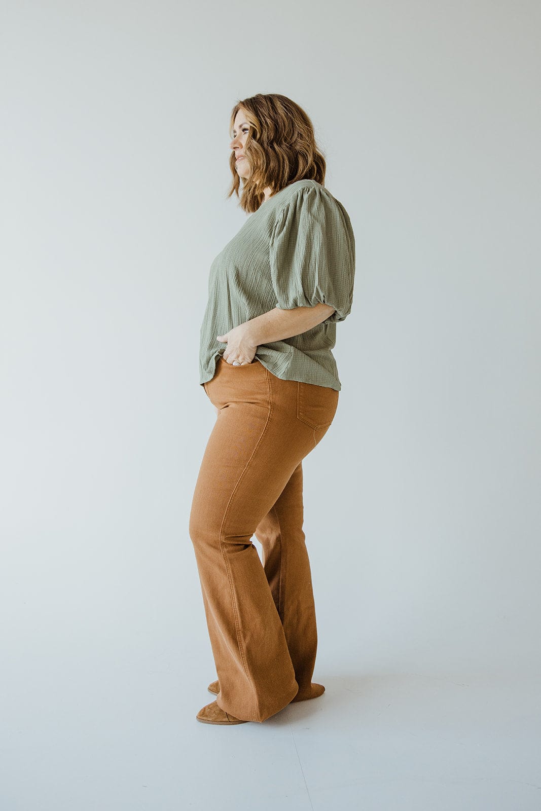 A woman stands sideways, wearing a green blouse with puffed sleeves and JUDY BLUE HIGH WAIST TUMMY CONTROL FLARE IN URBAN BRONZE pants by Judy Blue. She looks away with her hands in her pockets against a plain, light background.