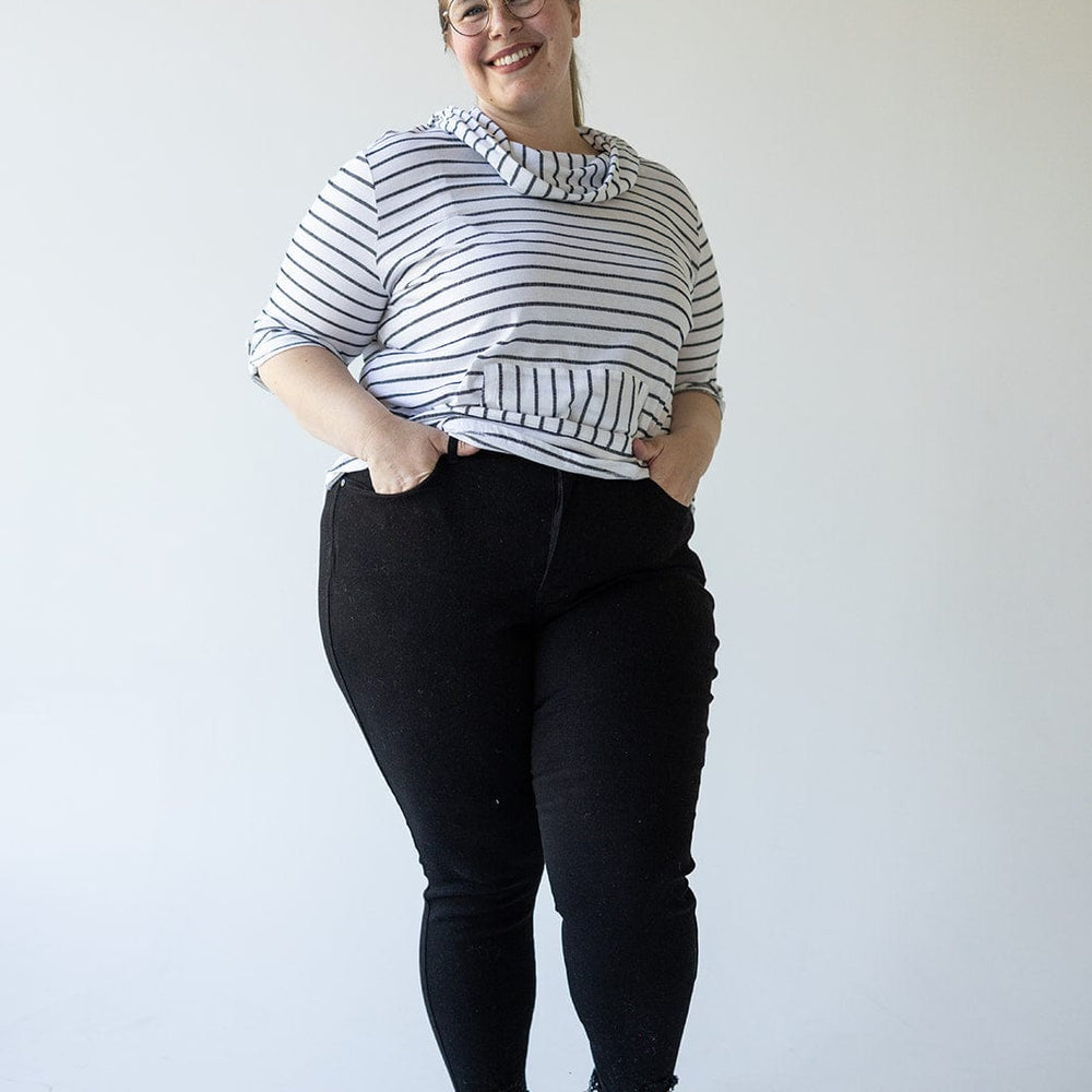 A person stands against a plain background, wearing a striped top, Judy Blue HIGH WAIST TUMMY CONTROL SKINNY JEANS WITH DISTRESSED HEM IN BLACK, and sandals, smiling with hands in pockets.